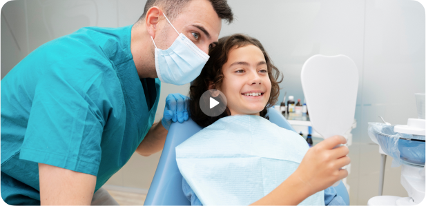 Friendly dentist smiling beside young boy examining his teeth in tooth-shaped mirror